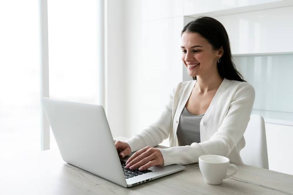 Mulher de cardigã branco digitando no notebook, com uma xícara ao lado